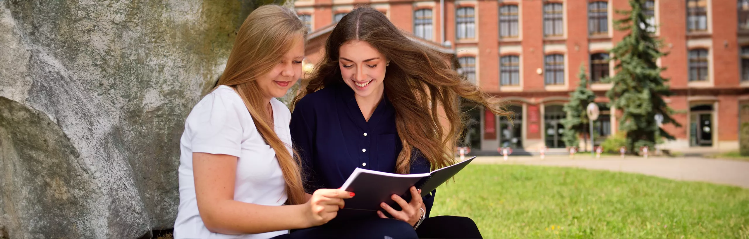 Studentki przeglądają notatki przed budynkiem uczelni.